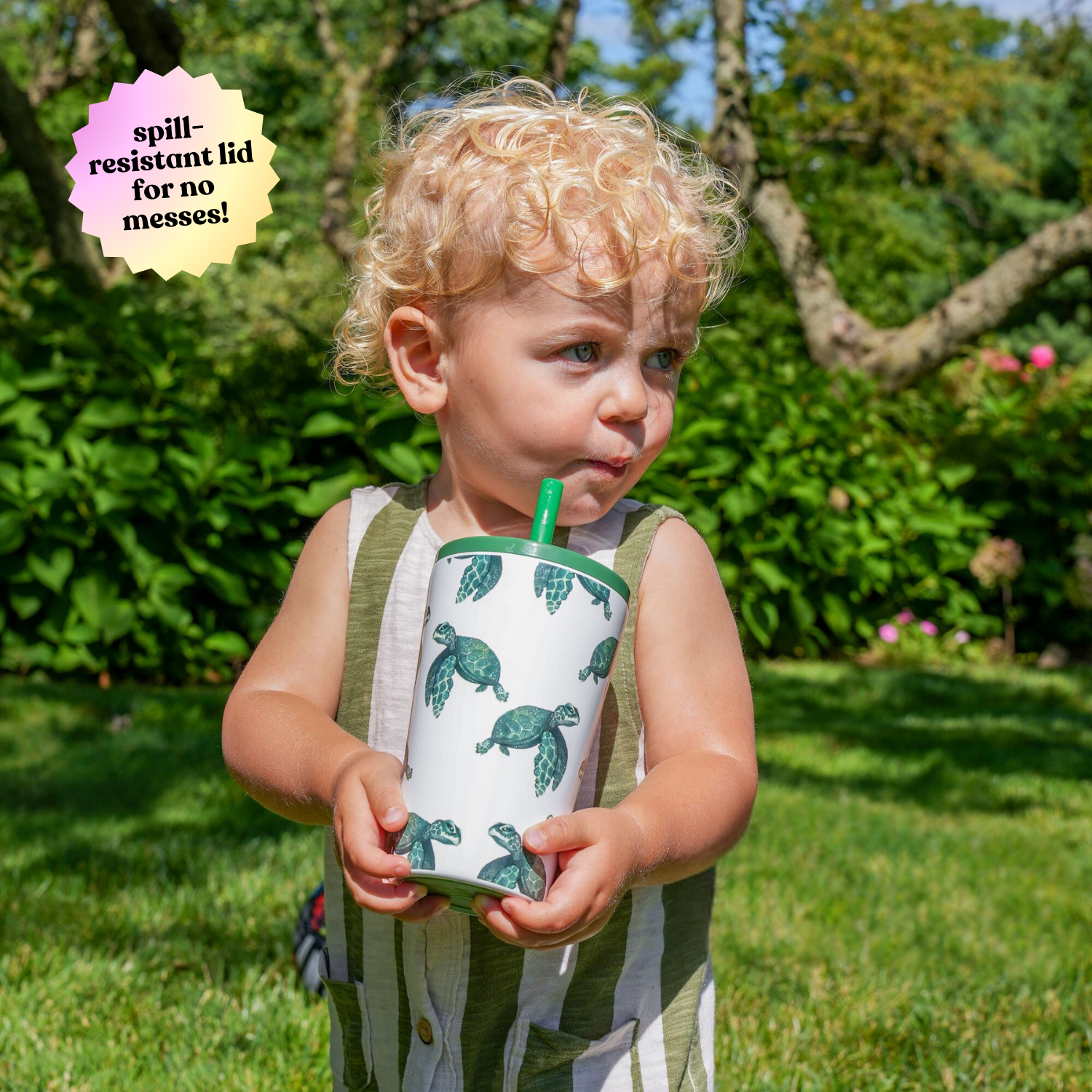 Young child outdoors drinking from a 14 oz stainless steel cup with a sea turtle design, featuring a built-in soft silicone straw and an inner stainless steel straw. The child is enjoying a sunny day in a lush green garden.