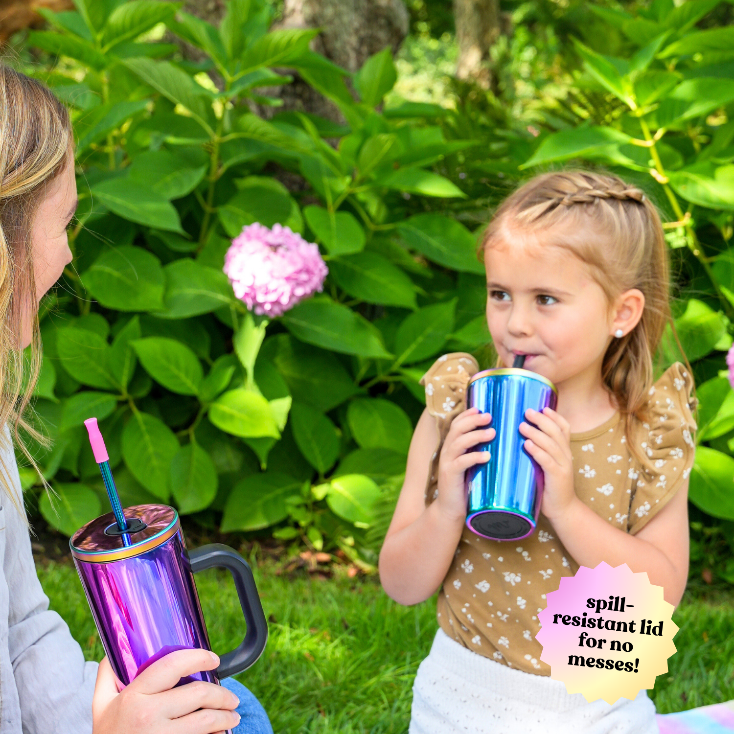 A mother and daughter sitting outside, each holding an iridescent stainless steel cup with a spill-resistant lid. The daughter drinks from her cup while the mother holds a similar cup with a handle. They are surrounded by lush green plants with a pink hydrangea in the background.