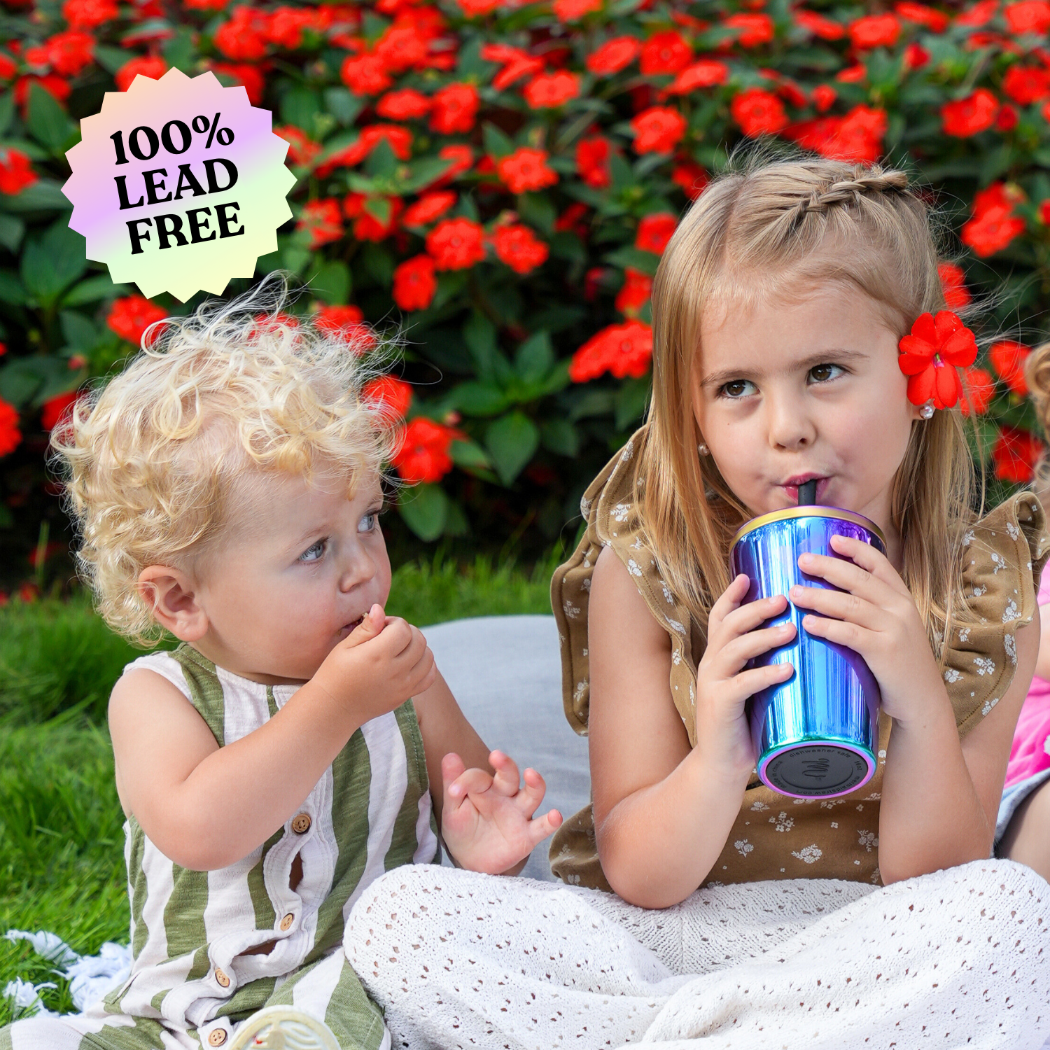 Two young children sitting on a blanket outdoors, with the girl drinking from an iridescent stainless steel cup and the boy holding a snack. The scene is set against a backdrop of vibrant red flowers, with a '100% lead-free' label displayed in the image.