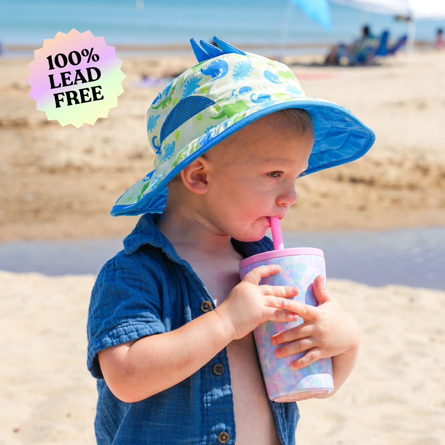 Young boy wearing a blue shirt and dinosaur sun hat, standing on a sandy beach while drinking from a pastel tie-dye 14 oz stainless steel cup with a pink straw. The image includes a '100% lead-free' label.