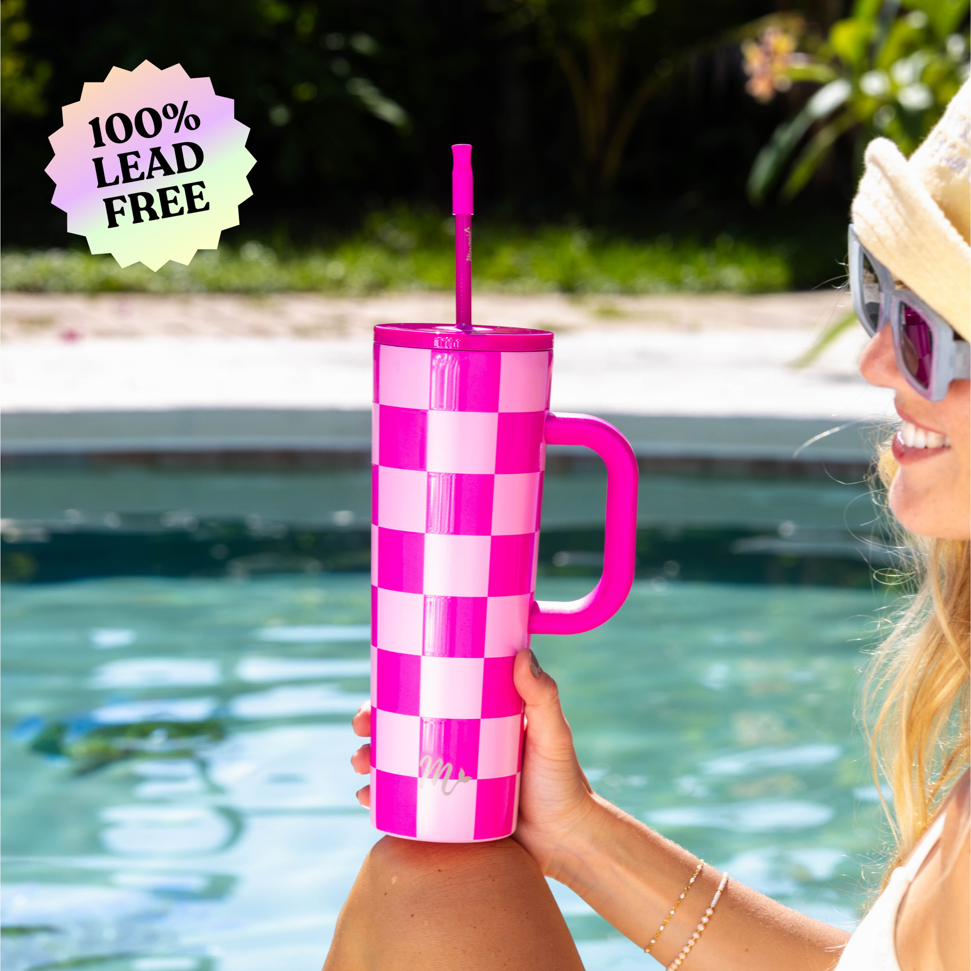 Woman sitting next to a pool holding a 30oz cup with a pink checkered design, pink handle, pink straw and pink silicone tip. The cup is labeled as 100% lead free