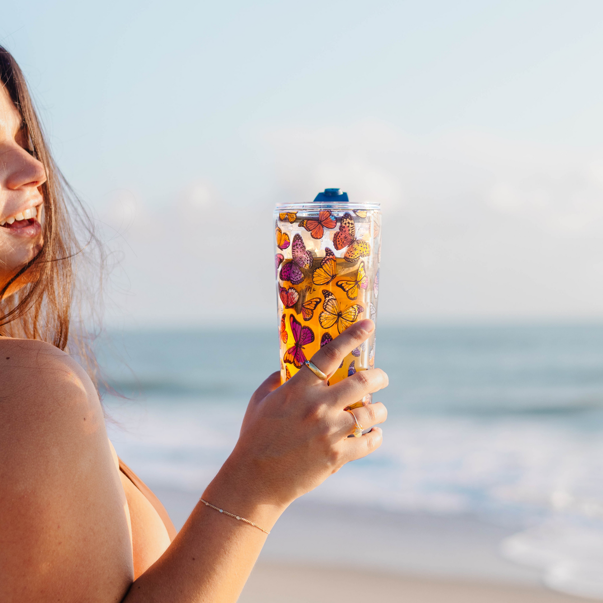 Person holding a 20oz double-wall glass cup with vibrant butterfly design, filled with iced tea, by the beach.