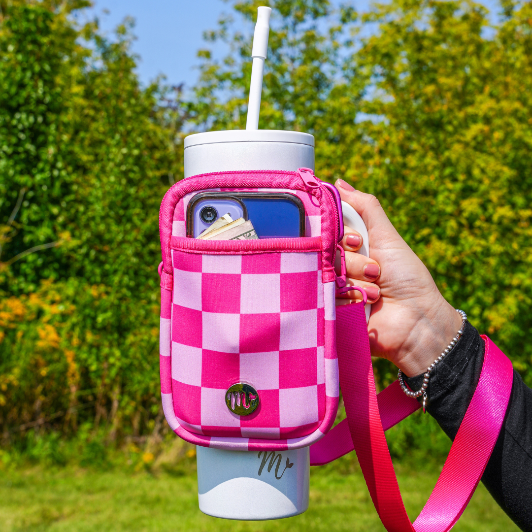 30 oz White Tumbler with Pink Checkered Sling - A hand holds the tumbler with a vibrant pink and light pink checkered sling, showcasing its convenient storage with a phone and cash visible in the front pocket. The adjustable strap allows for easy carrying, making it perfect for outdoor activities and on-the-go convenience.