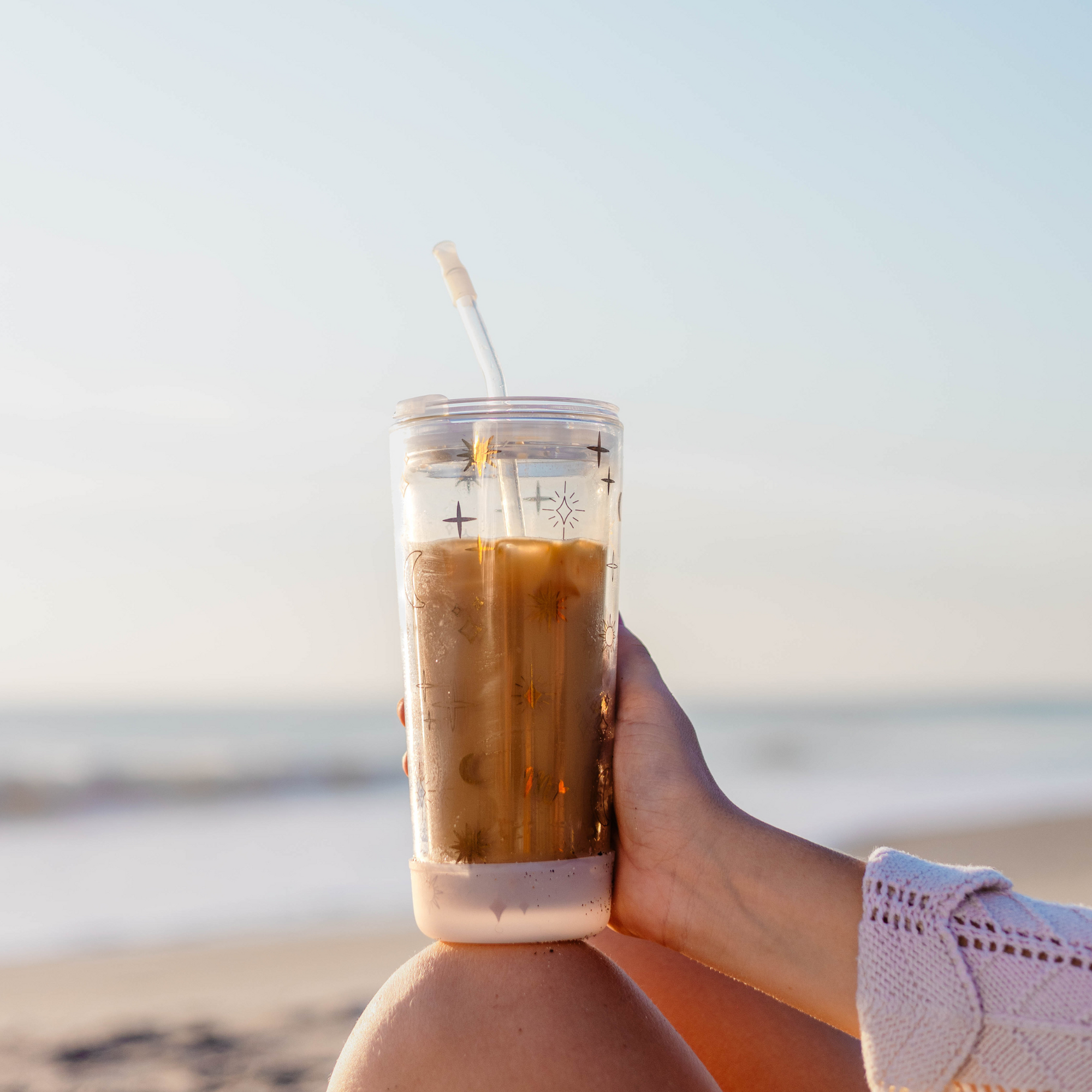 Person holding a 20oz double-wall glass cup with celestial design, filled with iced coffee, on a beach.