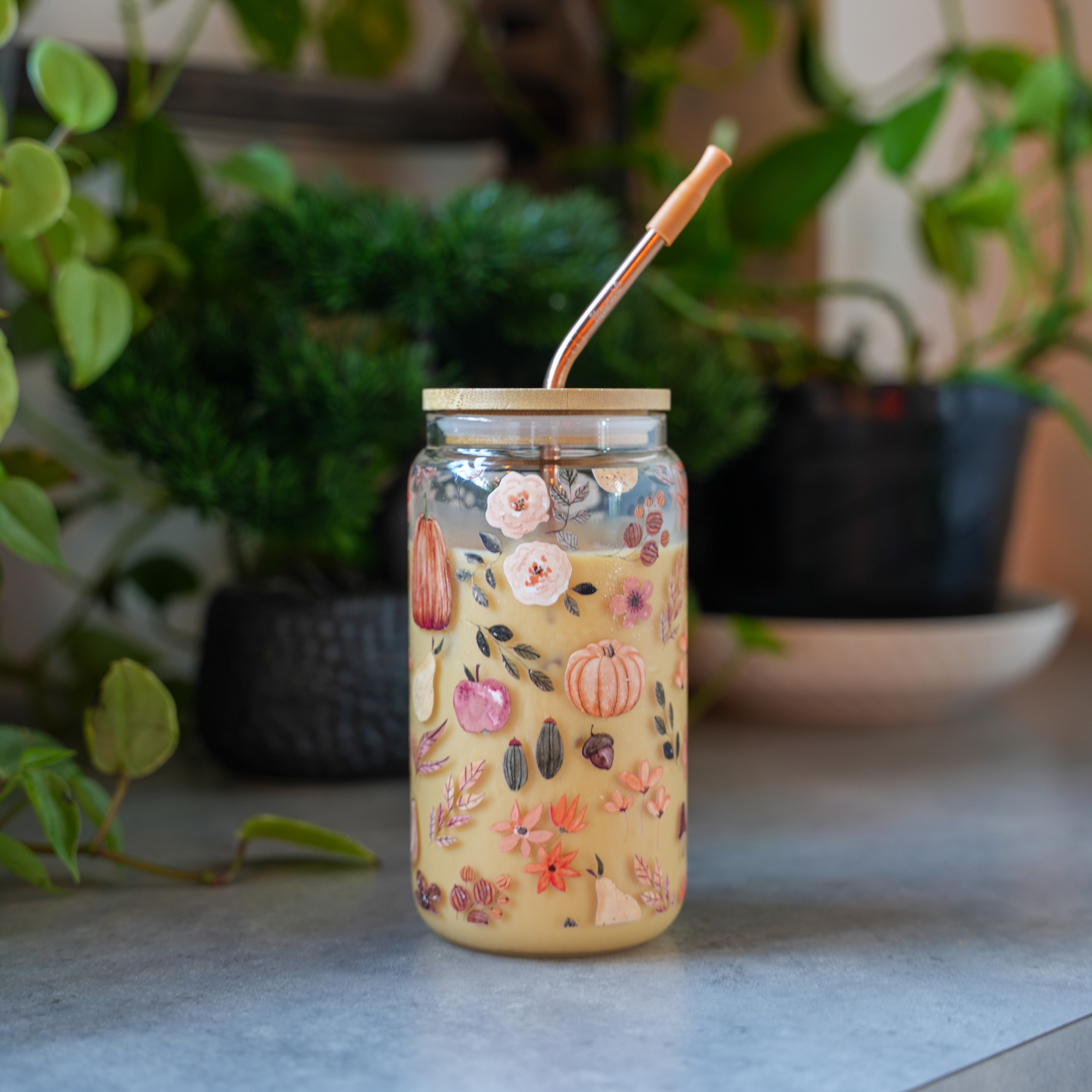 Autumn-themed glass tumbler with a wooden lid and reusable straw, placed on a table with greenery in the background.