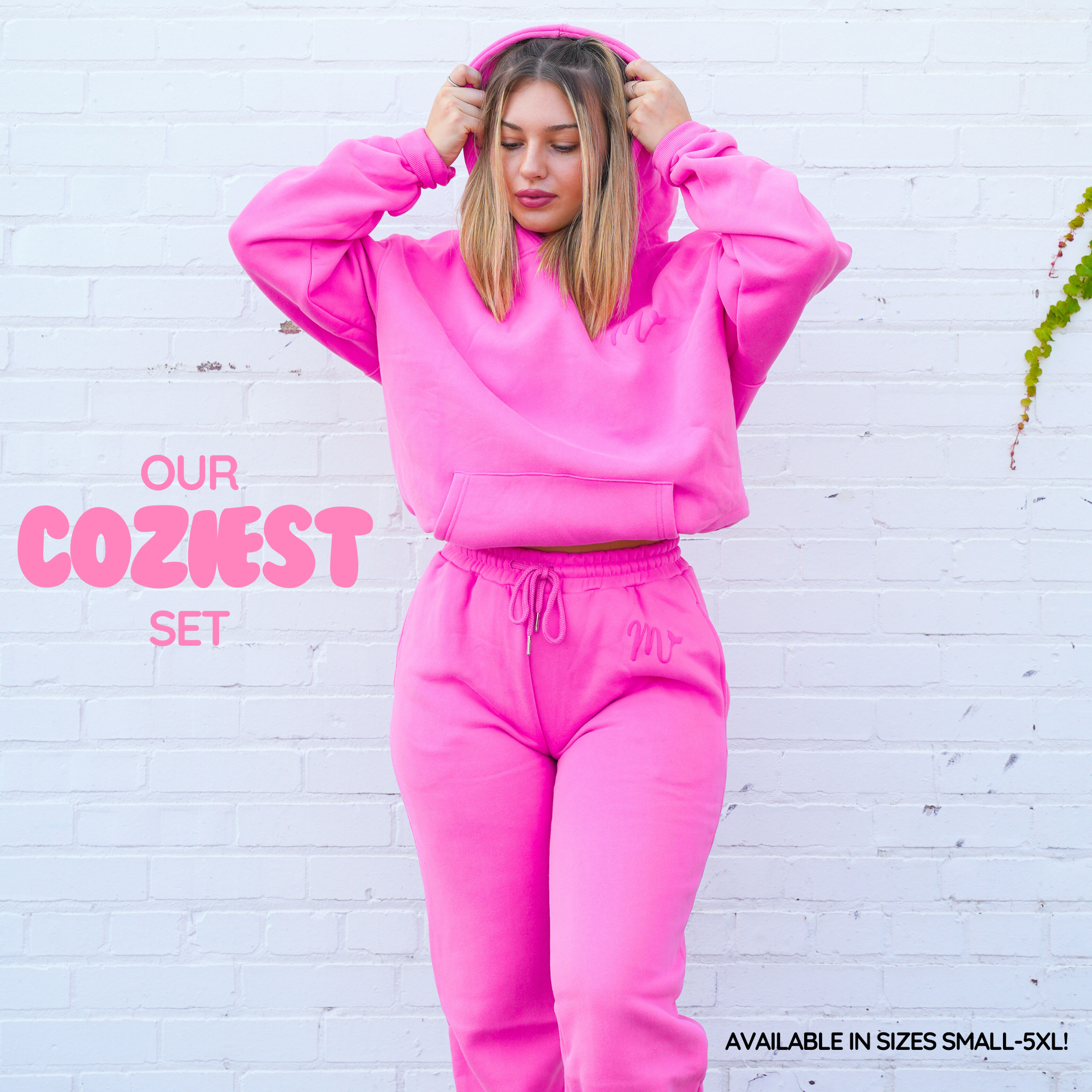 Model wearing a bright pink cozy hoodie and sweatpants set, showcasing a comfortable and stylish outfit. The hoodie features a subtle embossed logo, and the set is highlighted as 'Our Coziest Set,' available in sizes Small to 5XL. The model poses against a white brick wall, emphasizing the vibrant color and relaxed fit of the outfit.