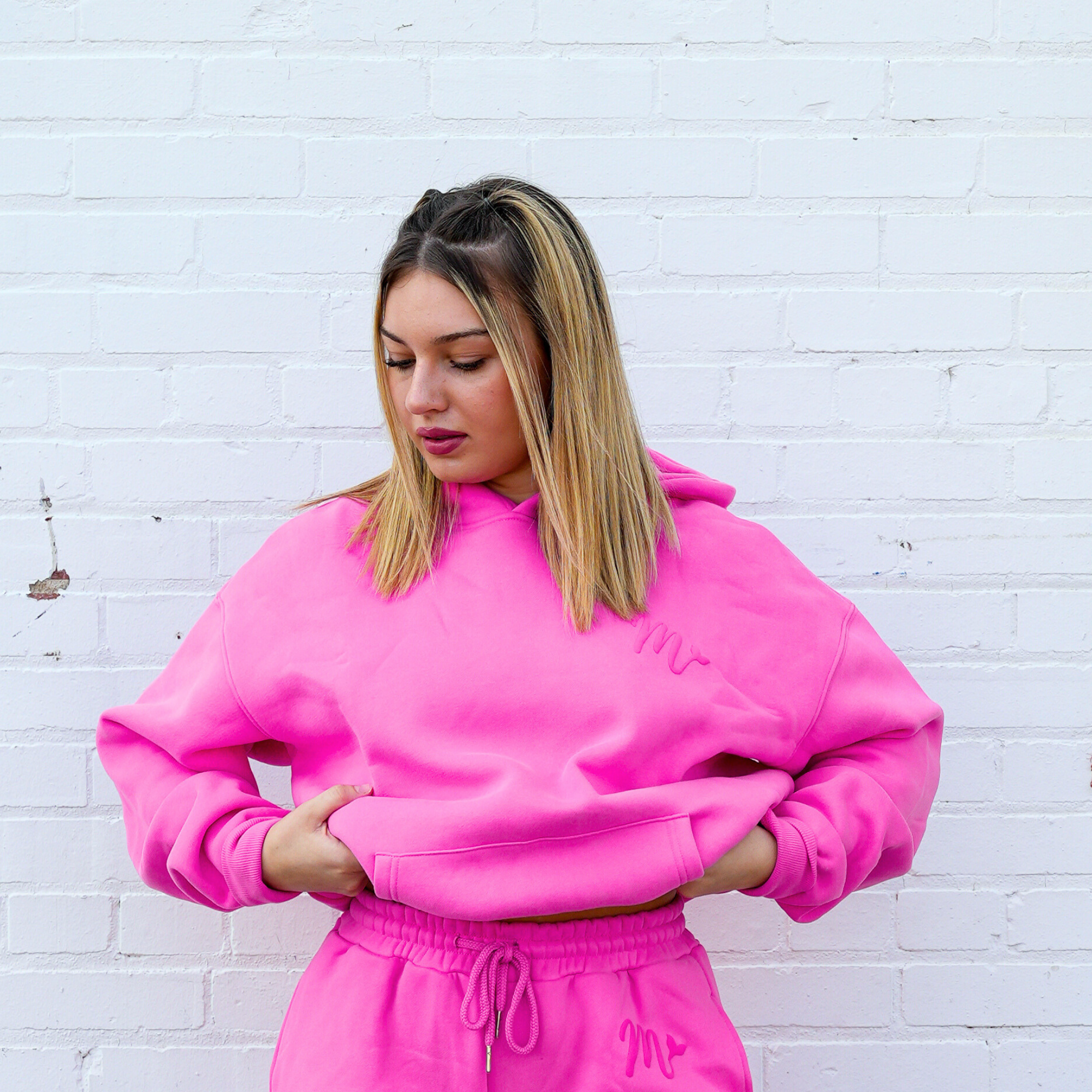 Model wearing a bright pink hoodie paired with matching sweatpants, both featuring an embossed logo. The hoodie has a relaxed fit, emphasizing comfort and style, as the model poses against a white brick wall, creating a vibrant and cozy look